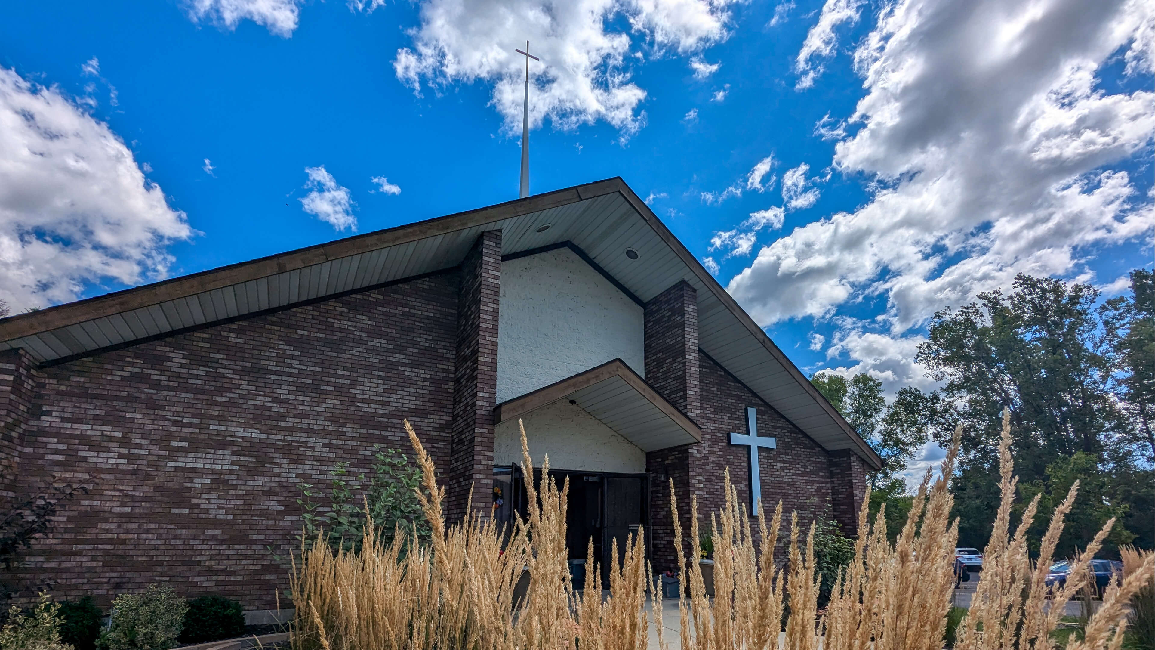 An exterior view of the front entryway to Lamb of God Lutheran Church. The stipel appears to pierce through the puffy white clouds on a beautiful summer day.